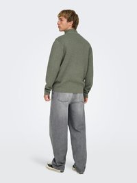 Pull en maille vert olive avec un col montant, associé à un jean denim gris ample et des baskets noires, vu de dos.