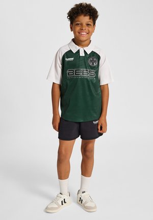 Zielono-biała koszulka polo z krótkim rękawem i kołnierzykiem, z napisem "BEES INTERNATIONALS" i logo; do tego czarne szorty i białe trampki.