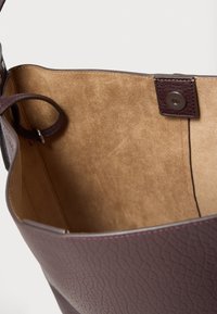 Interiorul unei genți din piele texturată maro, cu o căptușeală netedă bej. Are un nasture rotund maro cu capsa și o buclă laterală pentru atașare.