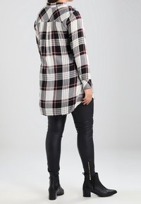 Lang skjorte i ternet mønster i sort, hvid og burgundy med et delt rygdesign. Kombineret med sorte leggings og ankelstøvler med gyldne lynlåse.