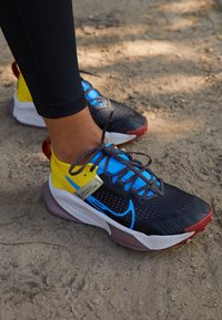 Chaussures de course Nike avec une tige en mesh noir agrémentée d'accents bleus et jaunes, un design texturé, et une semelle extérieure en caoutchouc rouge. Fermeture à lacets.