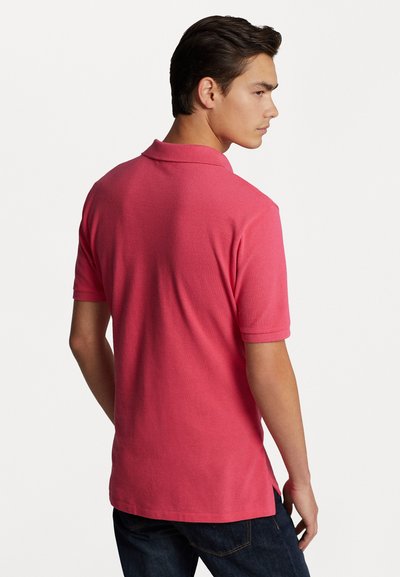 Polo shirt rose en tissu texturé, avec des manches courtes et un col. Le design comprend des fentes sur les côtés et un dos uni.