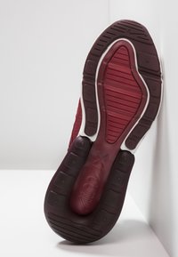 Sole tmavě červené a maroon tenisky Nike Air Max nakloněné proti bílé rohu, ukazující texturovaný vzor podrážky a logo.