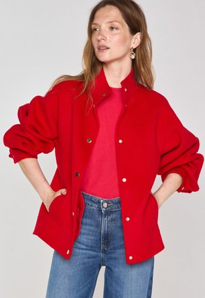 Giacca rossa, oversize, con colletto alto, grandi bottoni e maniche elasticizzate, abbinata a una camicia rossa e jeans blu.