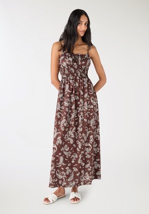 Kobieta w brązowej, długiej sukience maxi z białymi wzorami paisley oraz białych, płaskich sandałach typu slide, stojąca na tle jednolitego białego tła.