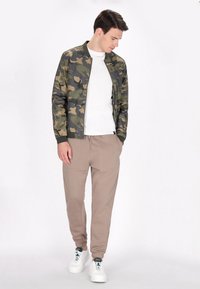 Camouflage bomberjack met geribbelde manchet, gecombineerd met beige joggers en witte sneakers. De outfit toont een ontspannen, stedelijke uitstraling.