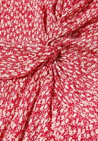 Rood patroon stof met een gerimpeld textuur, gekenmerkt door witte bloemmotieven. De stof is in het midden geknoopt, wat een geplooid effect creëert.