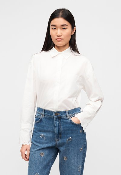 Λευκό πουκάμισο με μακριά μανίκια, γιακά με κουμπιά και τσέπη στο στήθος, συνδυασμένο με μπλε τζιν παντελόνι διακοσμημένο με λουλούδι κεντήματα.