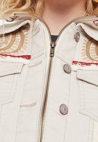 Primer plano de una chaqueta beige con cremallera y botones metálicos decorativos, motivos de sol bordados, ribete rojo y bordes deshilachados en los bolsillos.