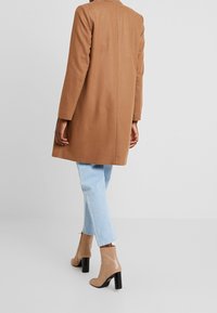 Brązowy wełniany płaszcz o prostym kroju, z długimi rękawami i detalami na tylnym szwie, zestawiony z jasnym denimu i beżowymi botkami za kostkę z czarnym obcasem.