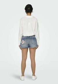 Shorts de mezclilla con dobladillo deshilachado, color azul desgastado y un detalle de parche en forma de diamante. Combinados con una camisa blanca ligera de manga larga.