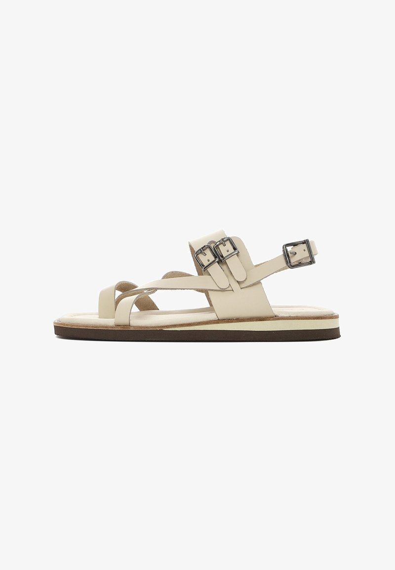 Beige leather sandal with multiple straps, featuring two adjustable metal buckles. Flat sole with a contrasting brown edge and soft insole.