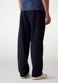 Marineblauwe wijde broek met contrasterende stiksels, voorzien van een ritszak aan de zijkant en een casual, ontspannen pasvorm. Gemaakt van zacht katoenen materiaal.