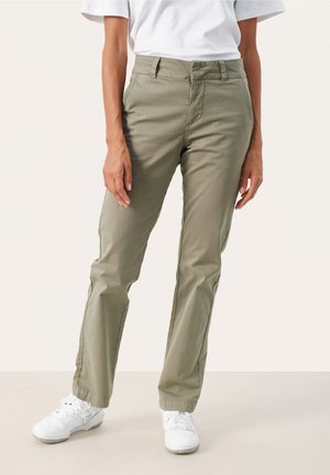 Femme portant un pantalon droit vert olive, des baskets blanches et une chemise blanche à manches courtes, debout devant un fond uni.