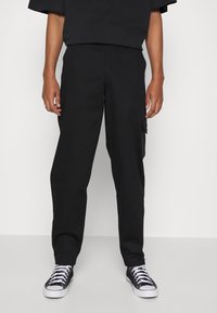 Pantaloni cargo neri con taglio dritto, tasca laterale con zip e tessuto liscio abbinati a sneakers nere alte.