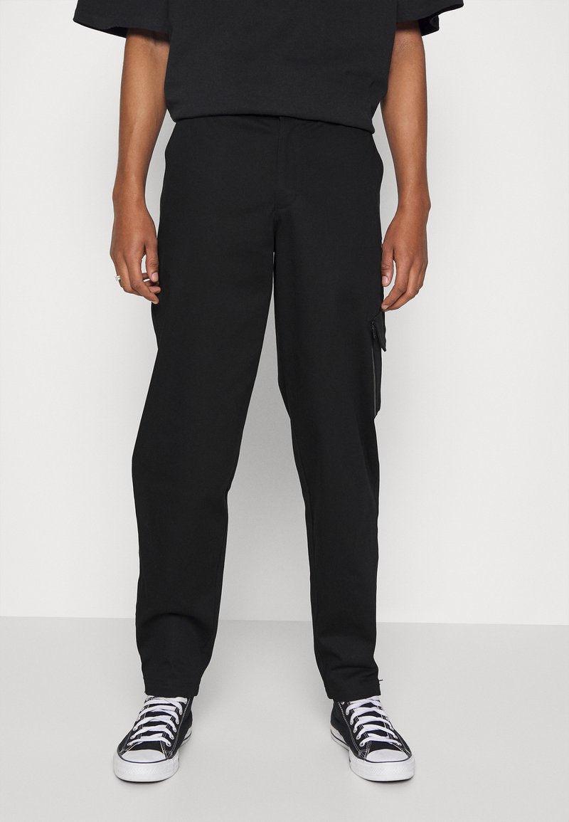 Pantaloni cargo neri con taglio dritto, tasca laterale con zip e tessuto liscio abbinati a sneakers nere alte.