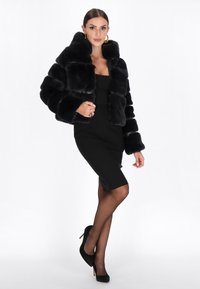 Sort faux fur cropped jakke med høj krave og boxy pasform. Parret med en tætsiddende sort kjole og sorte høje hæle. Glat tekstur.