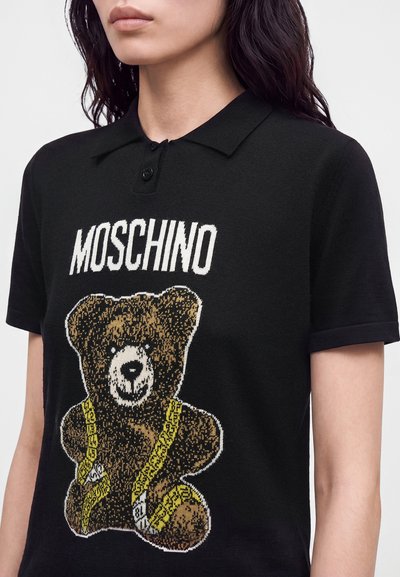 Femme portant un polo noir à manches courtes avec un dessin d'ours en peluche enroulé dans un ruban à mesurer jaune et le texte "MOSCHINO" au-dessus.