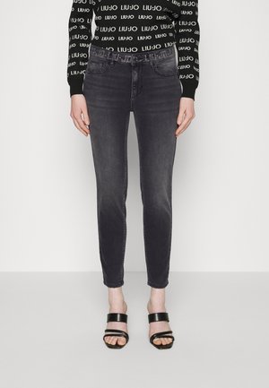 Kvinde iført sorte falmede skinny jeans med pyntet bælte og sorte remsandaletter med åben tå, stående mod en ensfarvet baggrund.