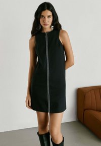 Jonge vrouw met lang donker haar, gekleed in een mouwloze zwarte jurk met een ritssluiting aan de voorkant en zwarte laarzen, staand binnen bij een bruine bank.