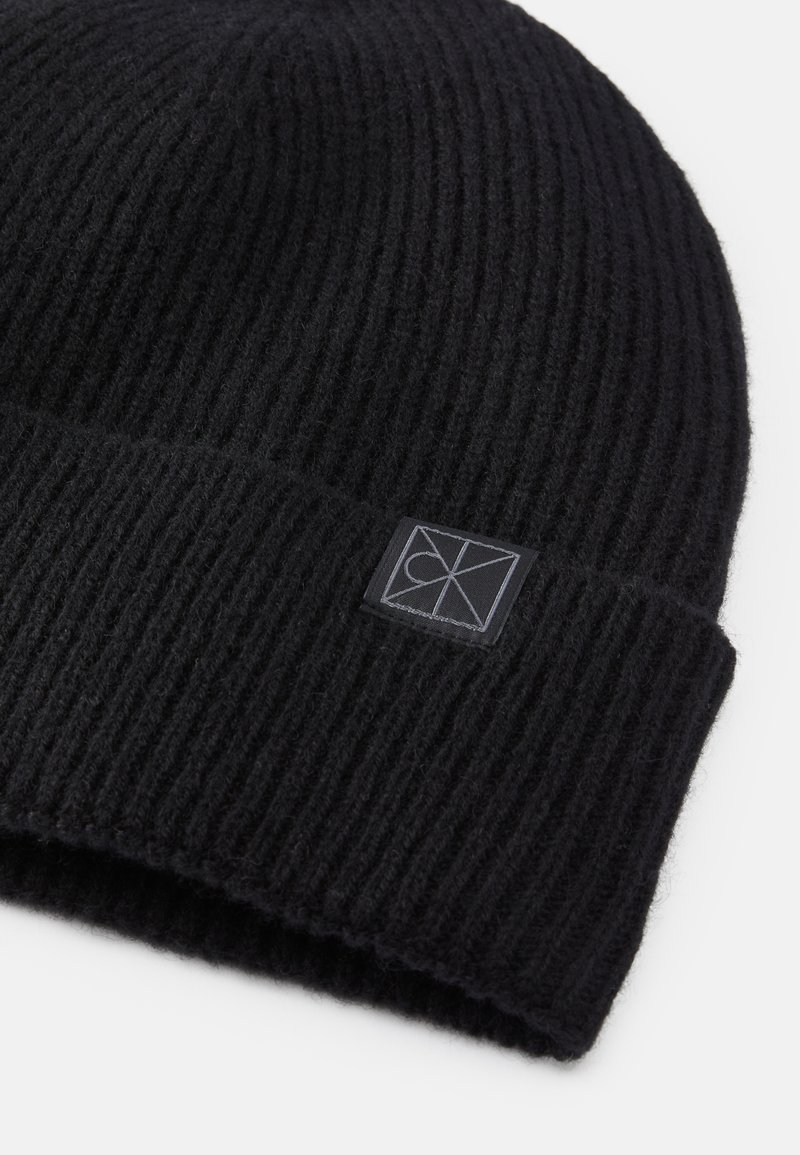 Sort strikket hue lavet af tekstureret uld, med en foldet kant og et lille sort logo-patch syet på forsiden.