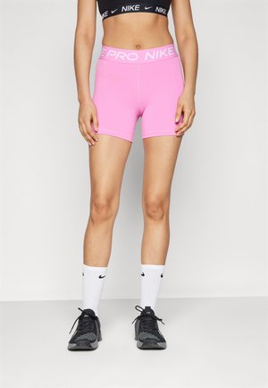Roosa Nike Pro lühikesed püksid valgete logoga vööga, koos mustade sportlikke kingade ja valgete sokkidega, millel on must swoosh.