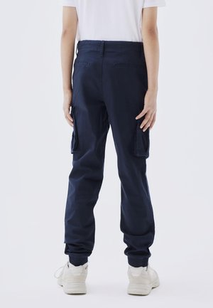 Persoon die marineblauwe cargobroek met elastische manchetten en witte sneakers draagt, met de rug naar de kijker toe, staand tegen een effen achtergrond.