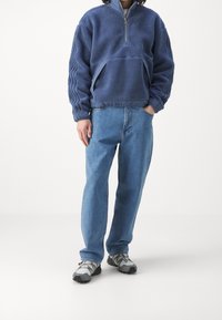 Blå fleecepullover med halvdragkedja, texturerad yta, sidfickor och elastiska ärmar, tillsammans med ljusblå jeans och grå sneakers.