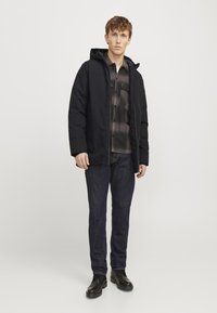 Sort puffers jakke med hætte, lagdelt over en grå ternet skjorte. Mørke jeans og sorte støvler fuldender outfittet. Glat tekstur hele vejen igennem.