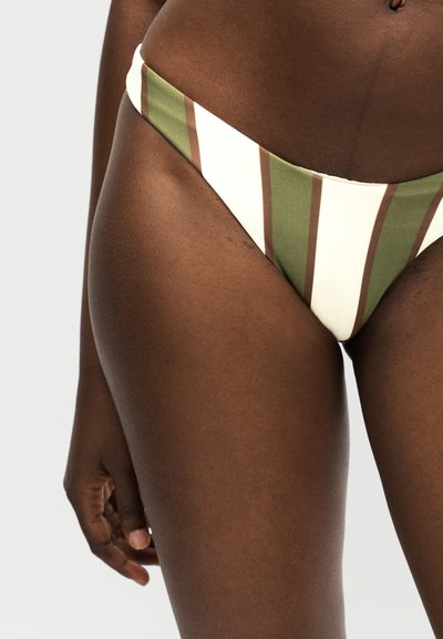 Κοντινό πλάνο ενός ατόμου που φοράει μαγιό με κάθετες πράσινες, λευκές και καφέ ρίγες, με το αριστερό χέρι χαλαρωμένο στο πλάι.