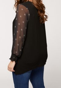 Black top with sheer, embroidered long sleeves featuring a floral pattern. Ruffled cuffs with elastic detail. Worn with blue jeans.