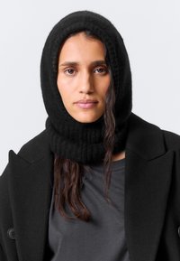 Gola alta de malha preta com design de capuz, apresentando uma textura canelada. Usada com um casaco preto por cima de uma camisa cinza escura.