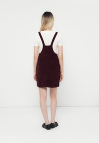 Robe salopette en velours côtelé bordeaux avec des bretelles réglables, une coupe droite et un ourlet fini, portée avec une chemise blanche en dessous.