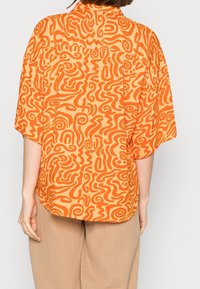 Oranje bedrukte shirt met abstracte krullen en bochten. Lichtgewicht stof, korte mouwen en een comfortabele pasvorm. Achteraanzicht weergegeven.