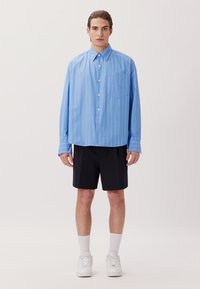 Camicia azzurro chiaro con righe verticali bianche, abbinata a pantaloni corti scuri. Il modello indossa sneakers bianche e calze di lunghezza media.