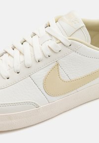 Bijele kožne tenisice s teksturiranim završetkom, bež Nike swoosh i ravnim bijelim vezicama. Imaju gumeni potplat s grip teksturom.