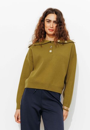 Un pull en maille vert olive avec un large col, trois boutons et un design court. Accompagné d'un pantalon sombre avec des rayures sur les côtés.