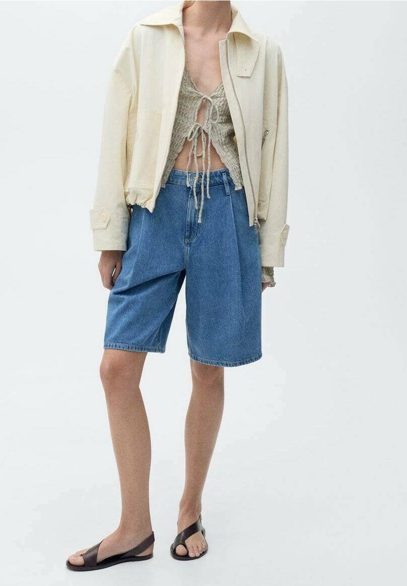 Personne portant une veste crème par-dessus un pull gris court noué à l'avant, un short en jean bleu d'une longueur au genou, et des sandales noires à bouts ouverts.
