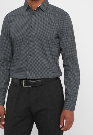 Mann trägt ein eng anliegendes dunkelgraues Langarmhemd mit Knöpfen und einem dezenten Rautenmuster, schwarzen Gürtel und dunkelgraue Hose.