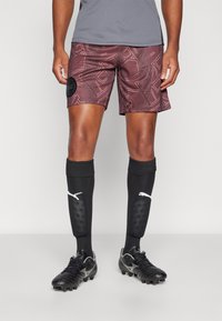 Mörkröda fotbollsshorts med geometrisk mönstring, svart logotyp-patch, tillsammans med svarta benskydd med vitt varumärke och svarta fotbollsskor.