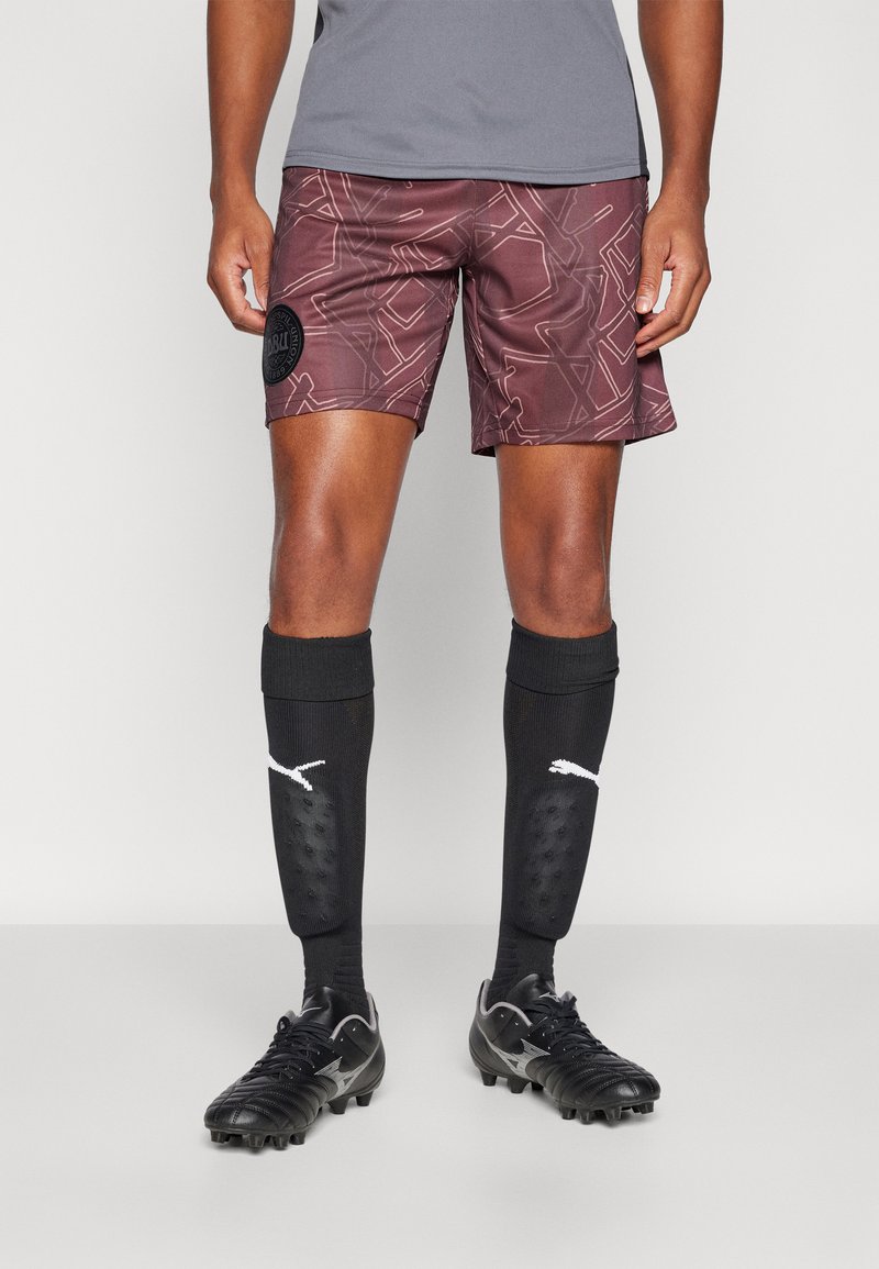 Osoba ubrana w bordowe, wzorzyste sportowe spodenki, czarne podkolanówki z białymi logo Puma oraz czarne korki, stojąca na szarej podłodze.