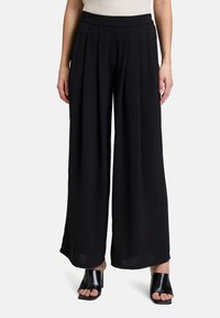 Zwarte wijde broek met een geplooid design, gemaakt van een lichtgewicht stof. Kenmerkt zich door een hoge taille en een zachte textuur. Gepaard met zwarte schoeisel.