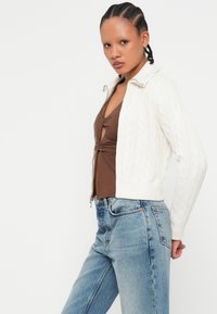 Gilet en tricot blanc avec fermeture éclair à l'avant, porté sur un haut marron et associé à un jean taille haute bleu clair. Tissu texturé et ourlet côtelé.