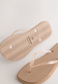 Tongs beiges avec semelles texturées et brides transparentes pailletées, dont une montrant une semelle à motif zigzag avec le nom de la marque embossé.