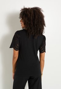 Blusa negra con mangas cortas perforadas y bordes en forma de pico, tejido texturizado y corte holgado. La vista trasera muestra un diseño sin adornos.