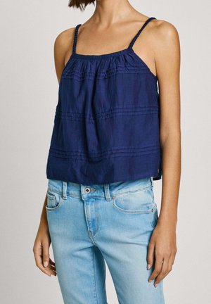 Femme portant un haut sans manches bleu marine à fines bretelles et un jean taille haute bleu clair, debout devant un fond uni.