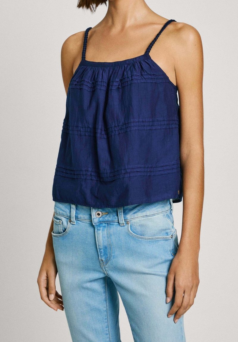 Femme portant un haut sans manches bleu marine à fines bretelles et un jean taille haute bleu clair, debout devant un fond uni.