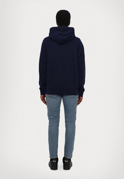 Sweatshirt à capuche bleu marine avec une coupe décontractée, des poignets et un ourlet côtelés, porté avec un jean bleu clair et des chaussures noires. Vue arrière affichée.