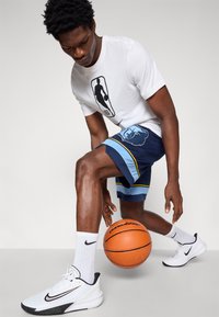 T-shirt branca com o logótipo da NBA em preto, calções azuis e azul-marinho com gráficos e riscas, sapatilhas brancas e pretas da Nike, e uma bola de basquete.