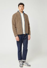 Giacca in pile marrone con colletto alto, dettagli blu e due tasche con zip, indossata sopra una camicia crema e abbinata a jeans in denim scuri.